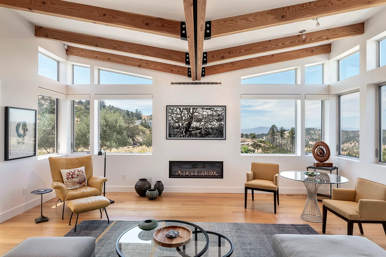 Modern living room with wood-beamed ceiling, large windows showing a scenic hillside view, a fireplace, leather and fabric chairs, and glass coffee tables on a hardwood floor.