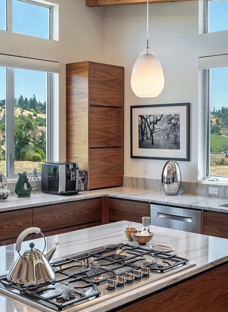 Modern kitchen with marble countertops, wooden cabinets, a built-in gas stove with a chrome kettle, and large windows showing a green landscape.