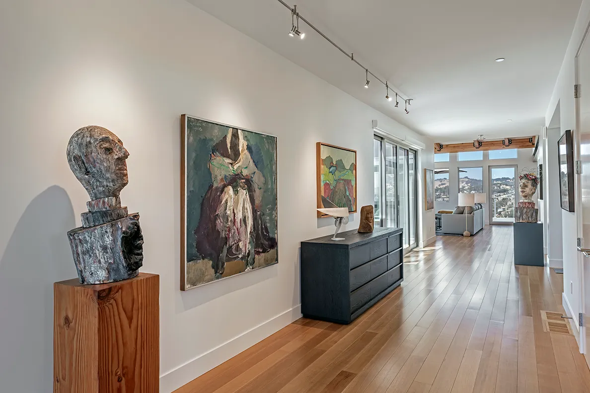 Modern hallway with wooden floor featuring abstract paintings and sculptures on pedestals and a black dresser, leading to a living room with panoramic windows showing a hillside view.