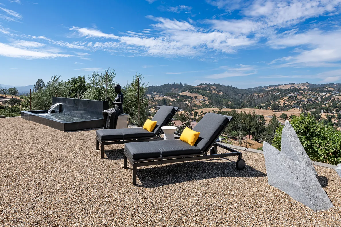 Outdoor lounge area with two gray cushioned chairs with yellow pillows, a small white side table, a black sculpture of a seated woman, a water fountain, and a mountainous landscape under a blue sky.