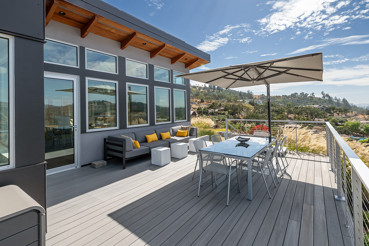 Modern outdoor deck with gray sofa and yellow cushions, glass dining table with chairs under large umbrella, overlooking scenic hillside landscape.