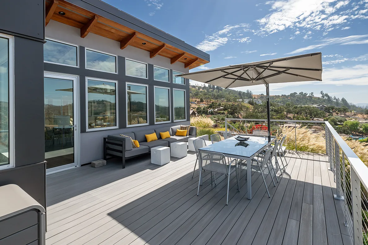 Modern outdoor deck with gray sofa and yellow cushions, glass dining table with chairs under large umbrella, overlooking scenic hillside landscape.
