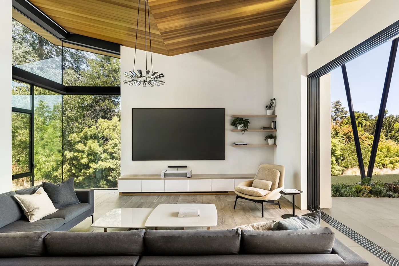 Modern living room with large floor-to-ceiling windows, a wall-mounted flat-screen TV, gray sofas, beige armchair, and wooden ceiling.