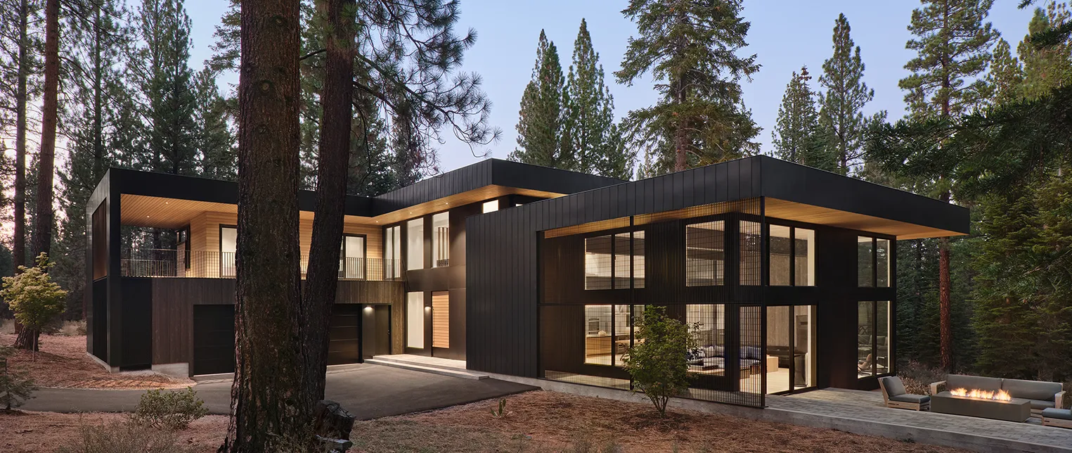 Modern black house with large windows, outdoor seating, and fire pit set in a pine forest at twilight.