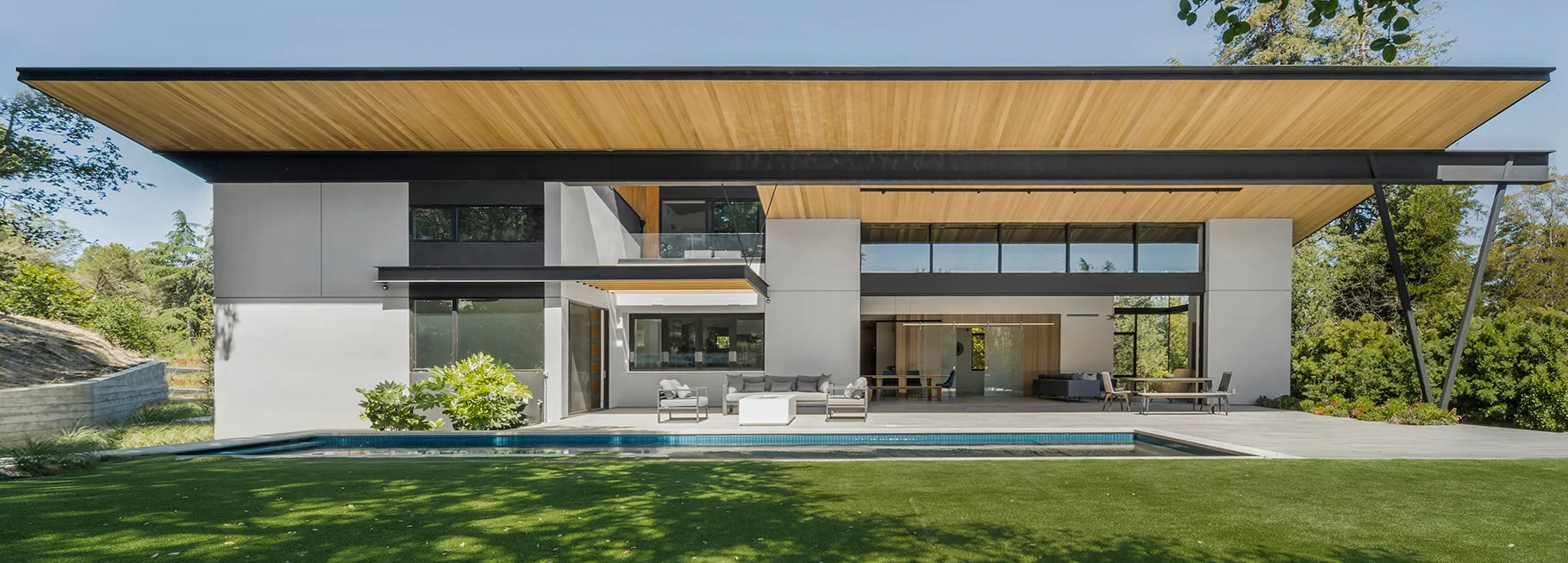 Modern wide exterior home with flat overhanging wooden roof, large windows, patio seating, swimming pool, and grassy yard.