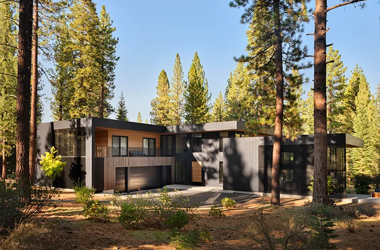 Modern two-story home with dark exterior and wooden accents surrounded by tall pine trees under a clear blue sky.