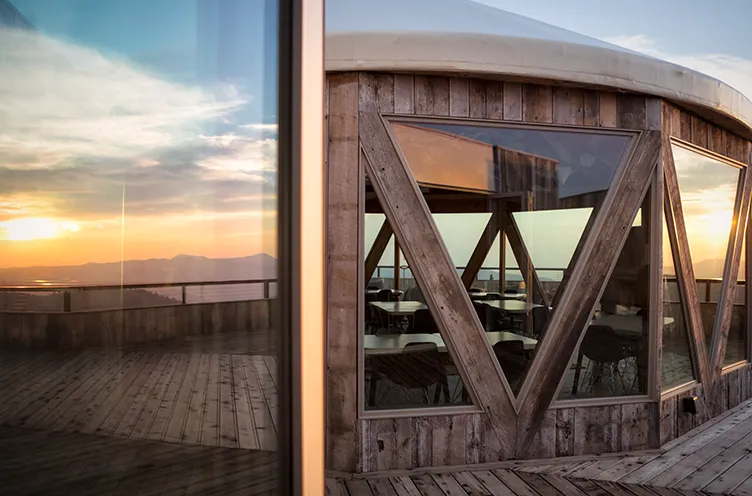 Wooden ski lodge with triangular windows reflecting sunset over mountains and a wooden deck.