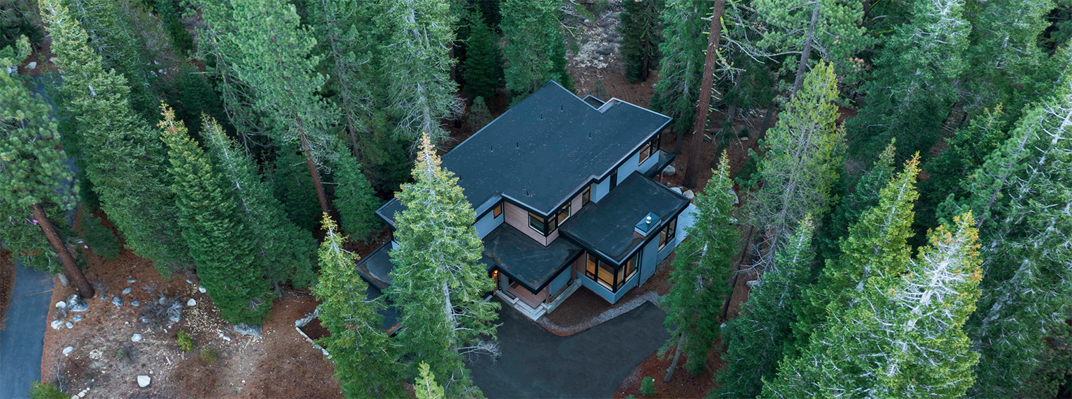 A modern two-story house with black roofs surrounded by tall evergreen trees in a forested area.