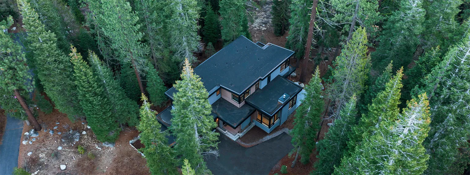 A modern two-story house with black roofs surrounded by tall evergreen trees in a forested area.