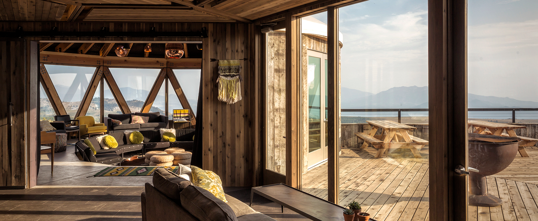 Interior view of a wooden lounge with large windows showing a seating area with colorful cushions and an outdoor deck with picnic tables and mountain views.