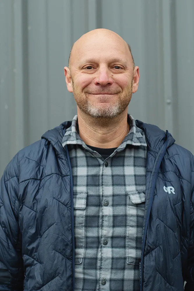 Smiling man with a short beard wearing a blue plaid shirt and a dark blue jacket standing in front of a gray metal wall.
