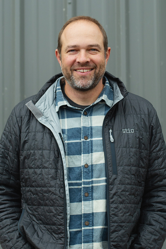Smiling man with short hair and beard wearing a blue and white plaid shirt under a black quilted jacket standing in front of a gray metal background.