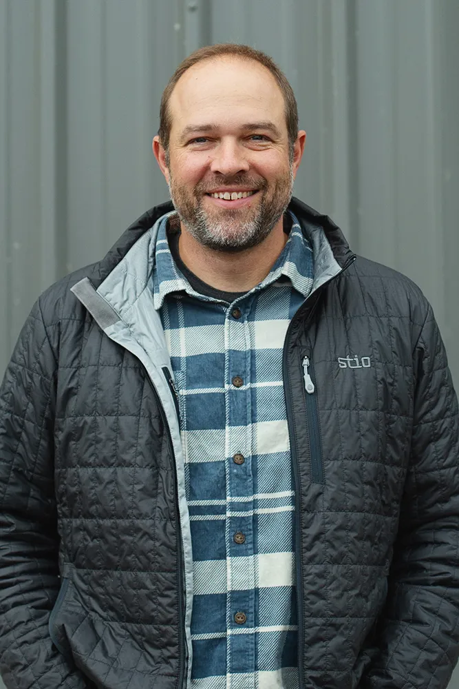 Smiling man with short hair and beard wearing a blue and white plaid shirt under a black quilted jacket standing in front of a gray metal background.