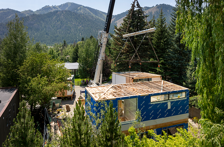 Crane lifting module above a partially built blue modular home surrounded by trees with mountains in the background.