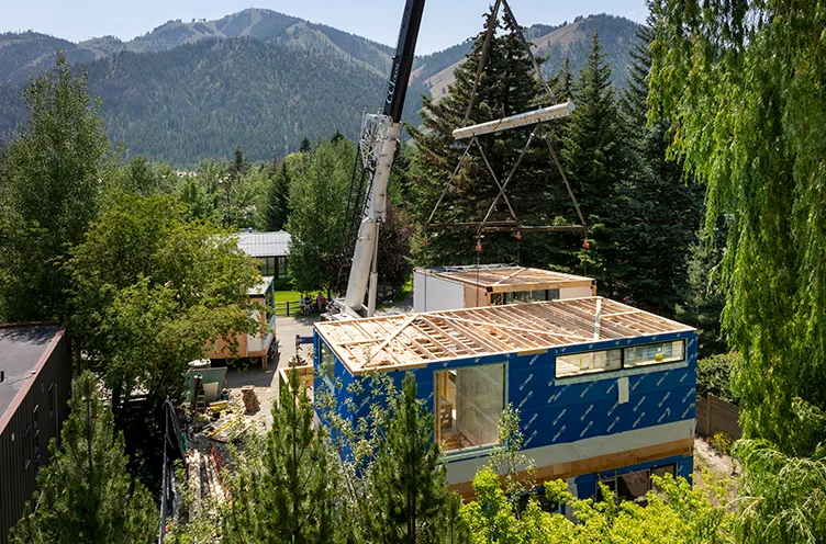 Crane lifting module above a partially built blue modular home surrounded by trees with mountains in the background.