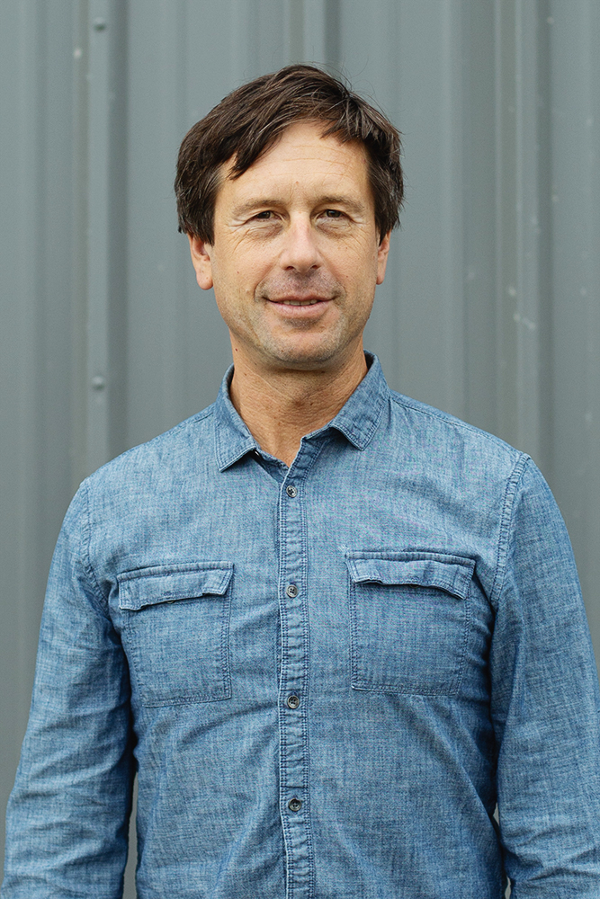 Man with short brown hair wearing a blue denim button-up shirt standing in front of a gray metal wall.