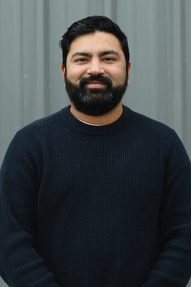 Smiling man with black hair and beard wearing a dark blue knitted sweater standing against a gray background.