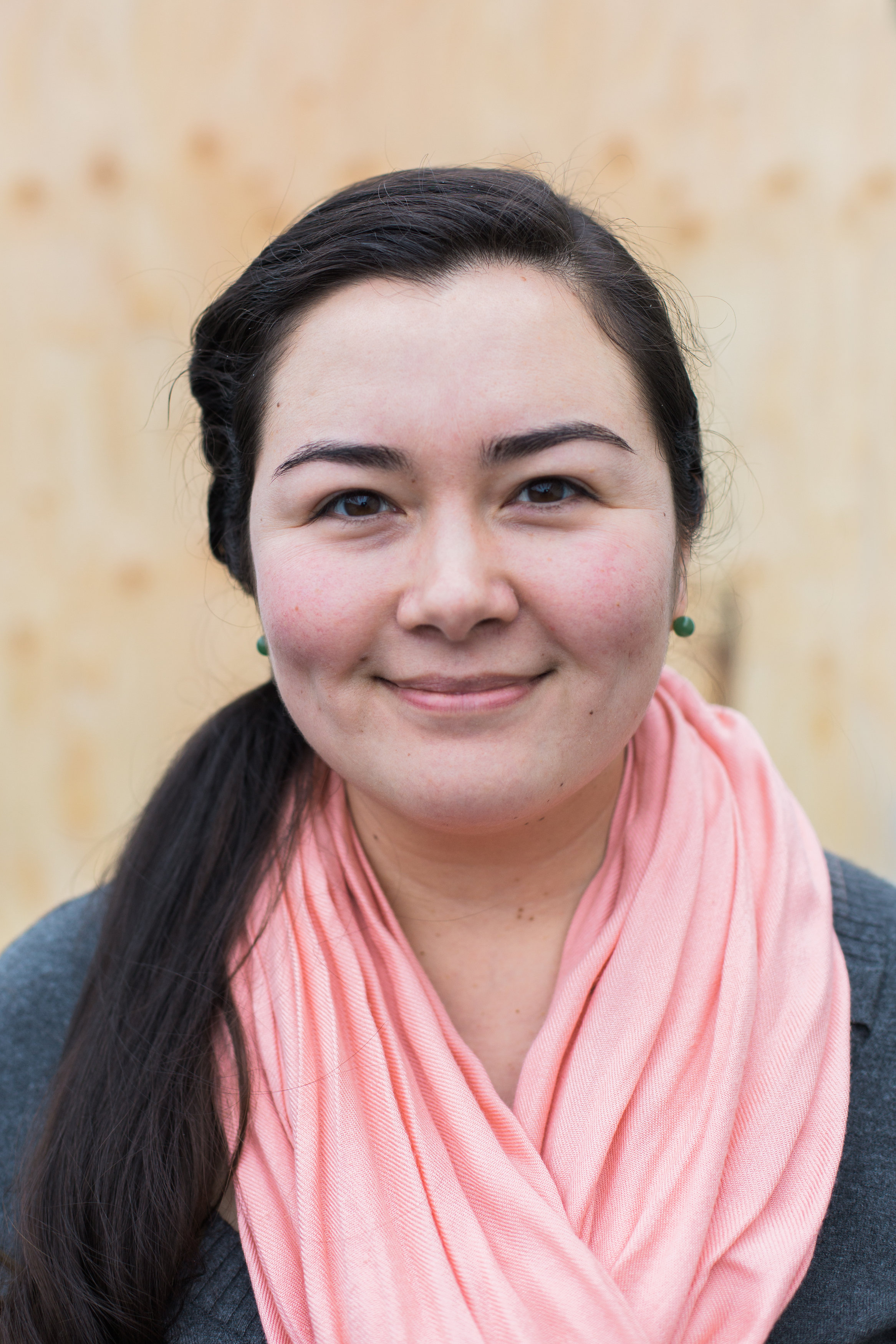 Smiling woman with long dark hair, pink scarf, and green earrings against a light wood background.