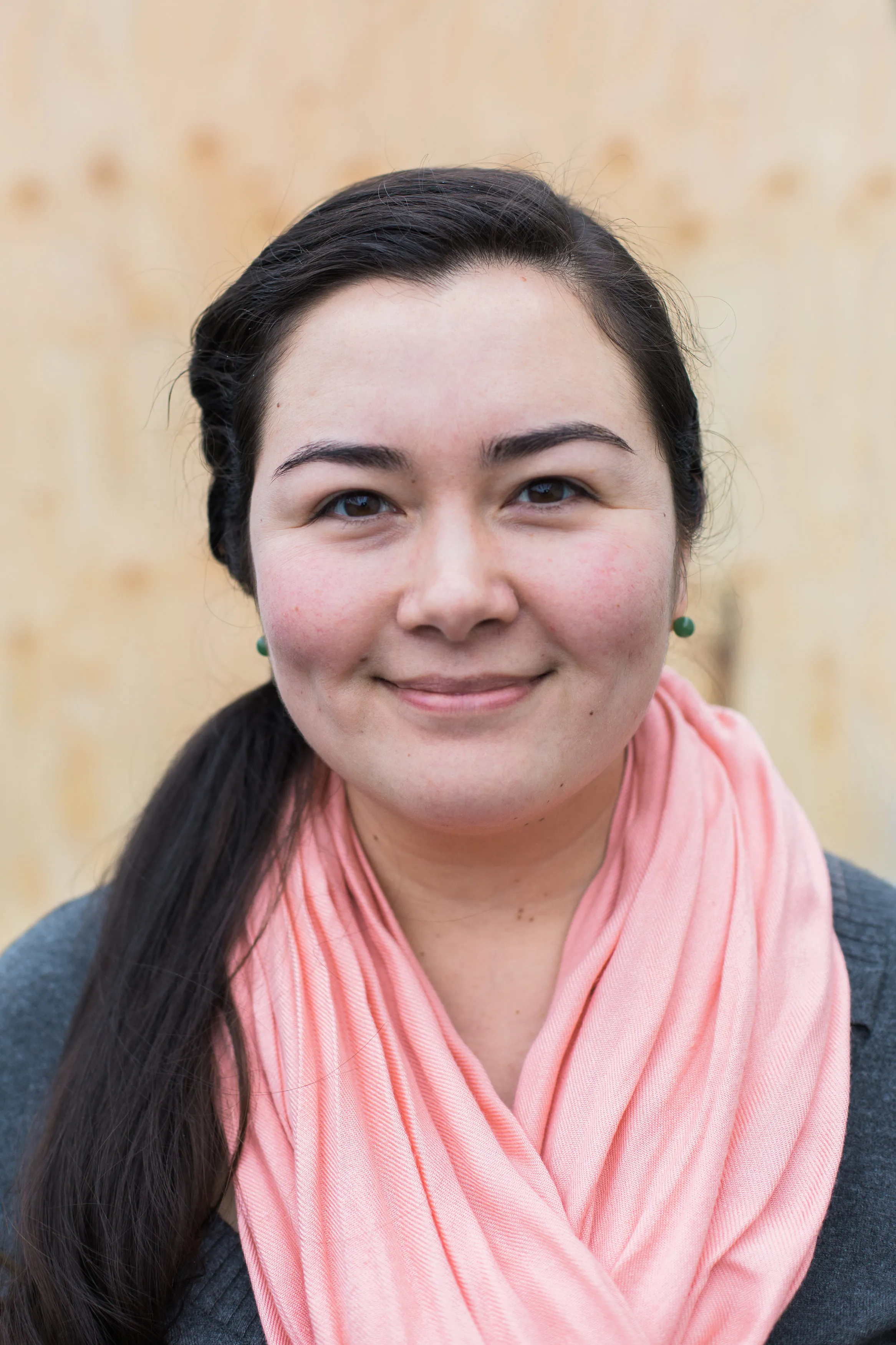 Smiling woman with long dark hair, pink scarf, and green earrings against a light wood background.