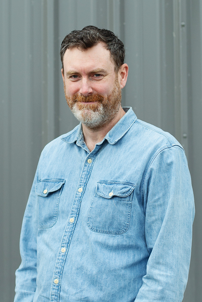 Man with dark hair and beard wearing a light blue denim button-up shirt standing against a gray metal background.