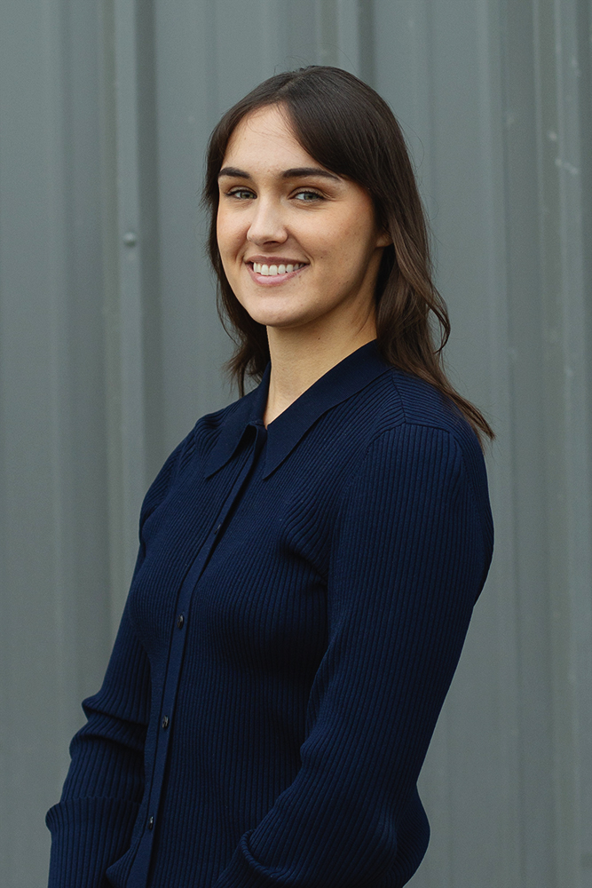 Smiling woman with medium-length brown hair wearing a navy blue ribbed sweater standing in front of a gray corrugated metal background.