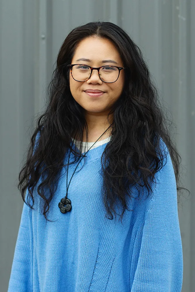 Woman with long black hair, glasses, and a blue sweater smiling against a gray background.