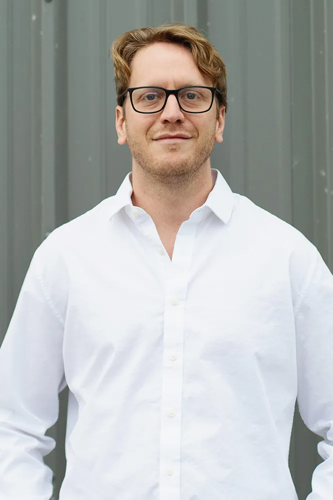 Man with light brown hair, glasses, and a white button-up shirt standing in front of a gray metal wall.