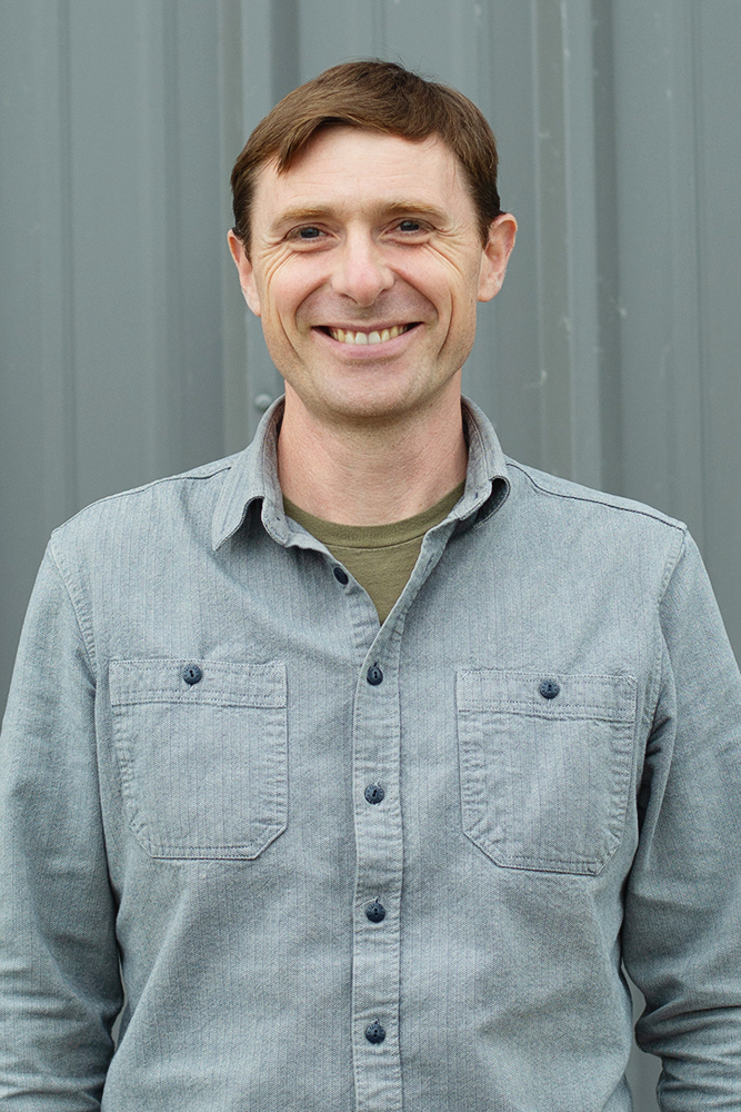 Smiling man with short brown hair wearing a grey button-up shirt standing in front of a grey background.