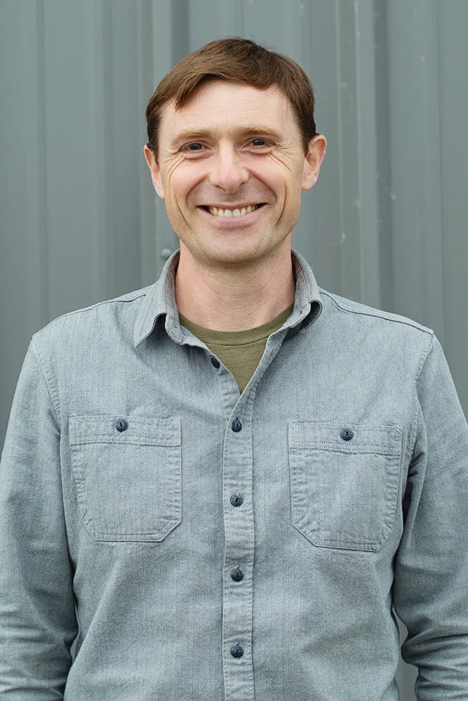 Smiling man with short brown hair wearing a grey button-up shirt standing in front of a grey background.