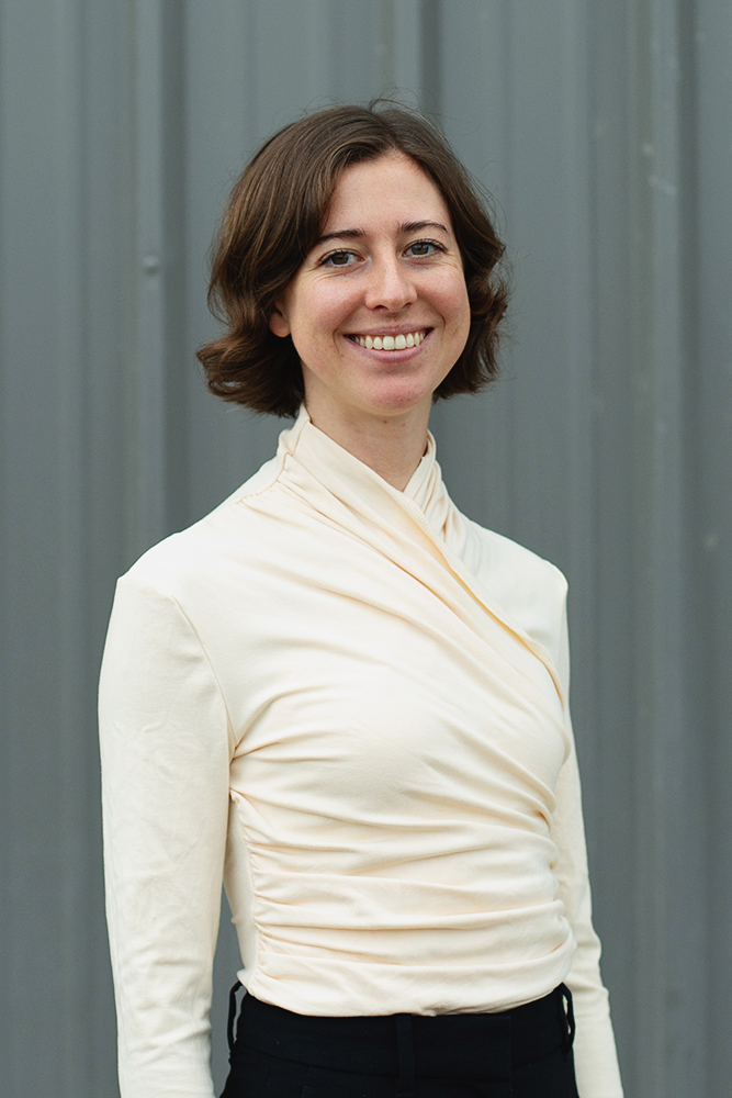 Smiling woman with short brown hair wearing a light cream-colored long-sleeve top against a gray metal background.