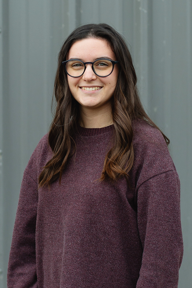 Smiling young woman with long brown hair and glasses wearing a purple sweater against a gray background.