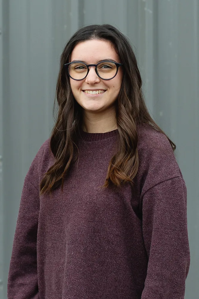 Smiling young woman with long brown hair and glasses wearing a purple sweater against a gray background.