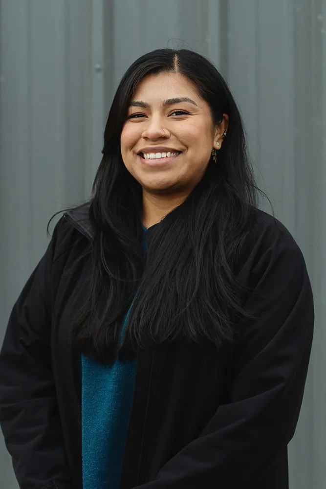 Smiling woman with long dark hair wearing a black jacket and teal sweater standing in front of a gray wall.