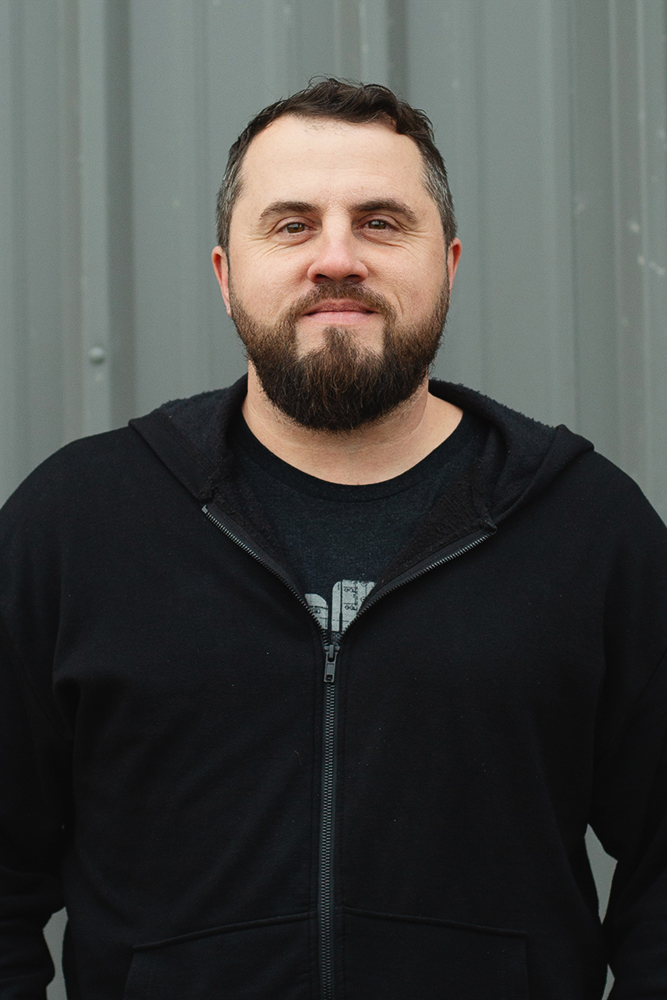 Bearded man wearing a black zip-up hoodie standing in front of a gray corrugated metal wall.
