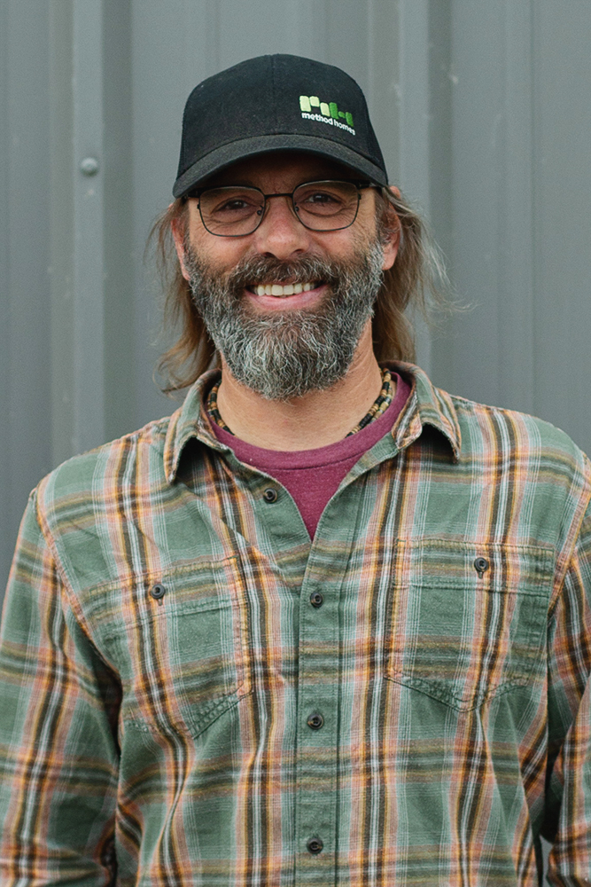Smiling man with glasses, a beard, and long hair wearing a black Method Homes cap and a green plaid shirt.