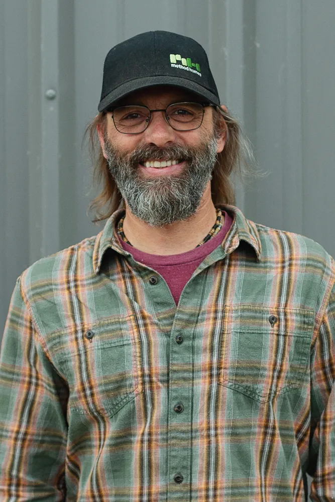 Smiling man with glasses, a beard, and long hair wearing a black Method Homes cap and a green plaid shirt.