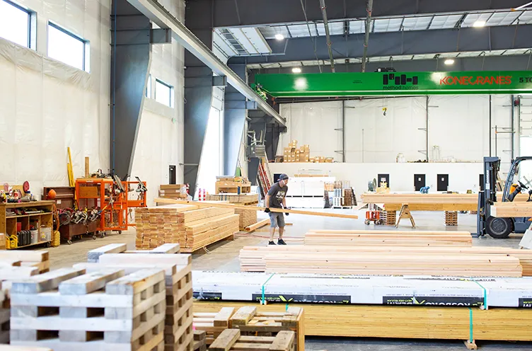 Worker moving long wooden planks inside a spacious, well-lit lumber warehouse.