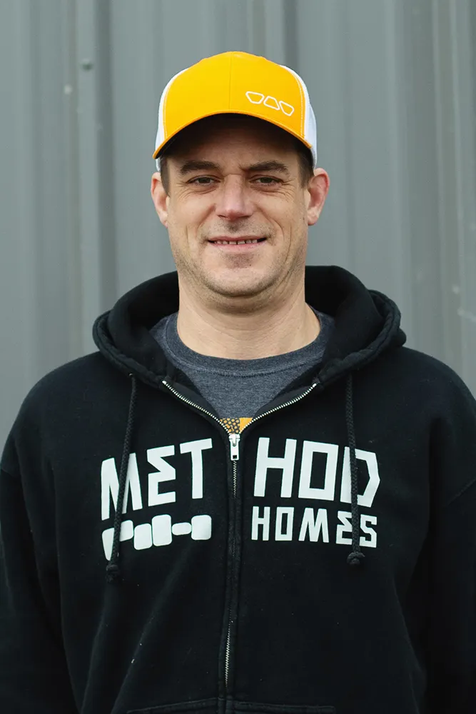 Smiling man with glasses, a beard, and long hair wearing a black Method Homes cap and a green plaid shirt.