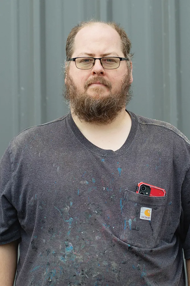 Smiling man with glasses, a beard, and long hair wearing a black Method Homes cap and a green plaid shirt.