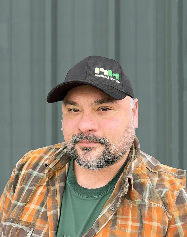 Smiling man with glasses, a beard, and long hair wearing a black Method Homes cap and a green plaid shirt.