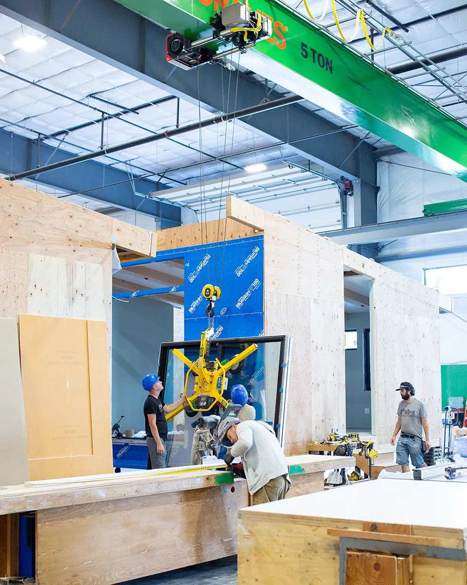 Workers installing a large window using an overhead crane inside a factory workshop with plywood walls and construction materials.