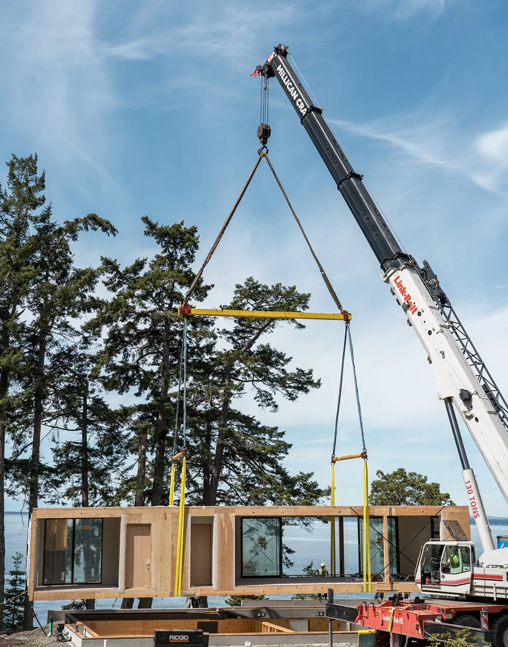 Crane lifting a large wooden modular home section above a construction foundation near trees and water.
