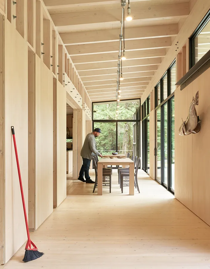 Man arranging items on a wooden dining table inside a modern room with large windows and wooden beams.