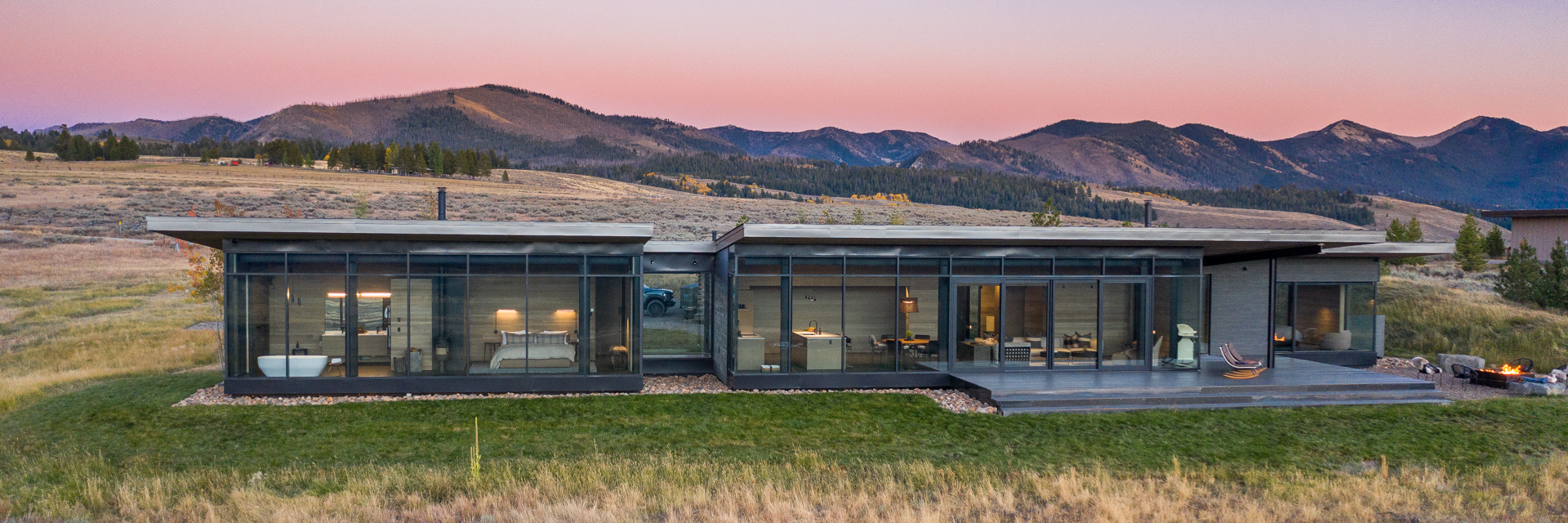 Modern glass-fronted home with mountain backdrop at sunset and outdoor fire pit on patio.