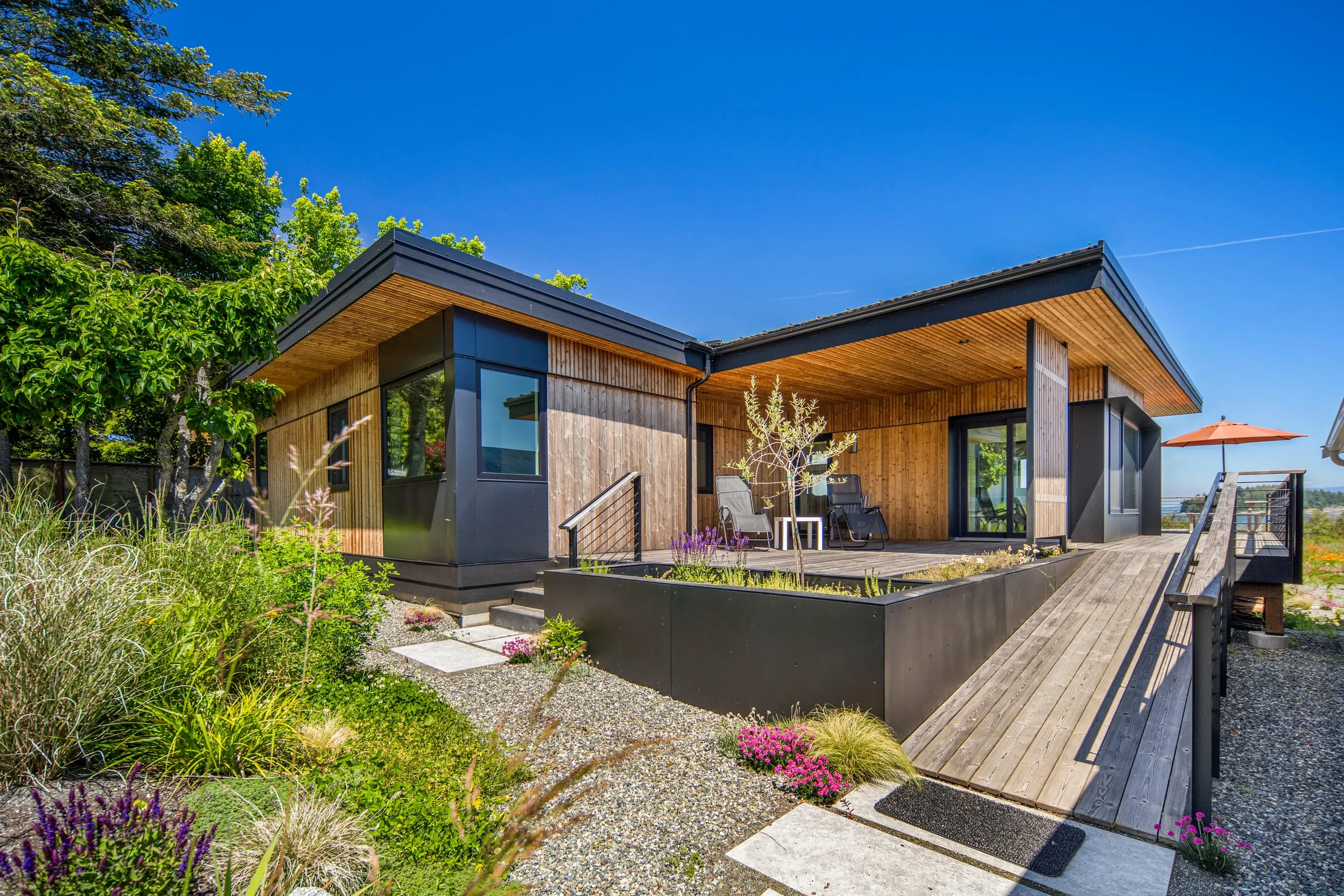 Modern single-story house with wooden exterior, large windows, a front porch, and a garden with greenery and rocks under a clear blue sky.