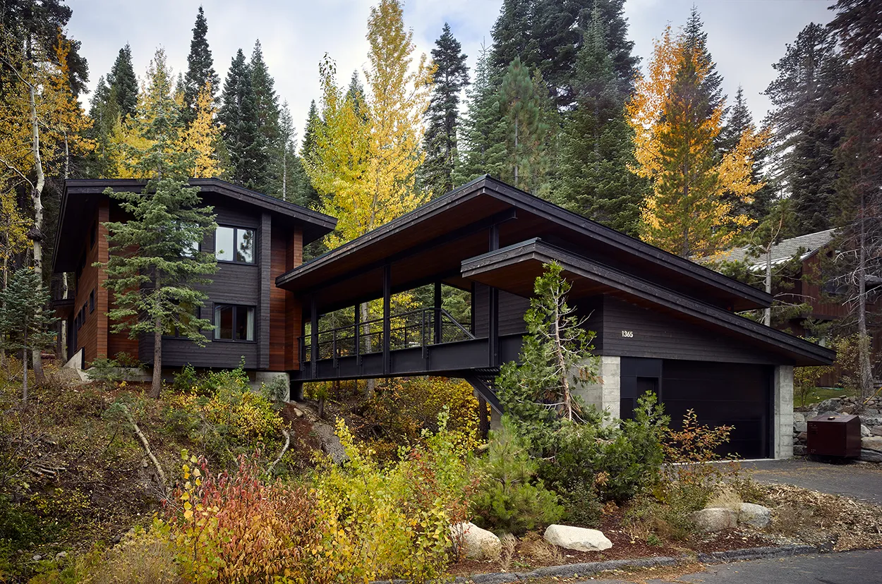 Modern dark wood house with carport surrounded by autumn trees and dense forest greenery.
