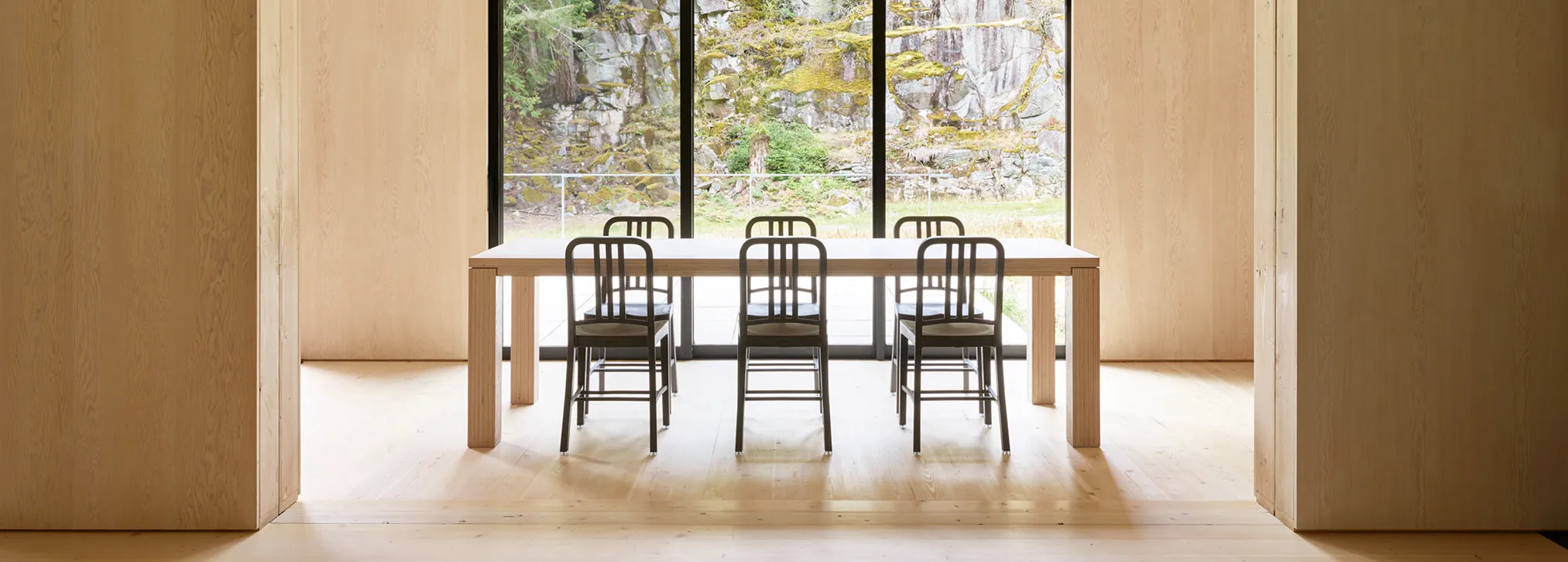 Minimalist dining area with six black chairs around a wooden table by large glass doors overlooking rocks.