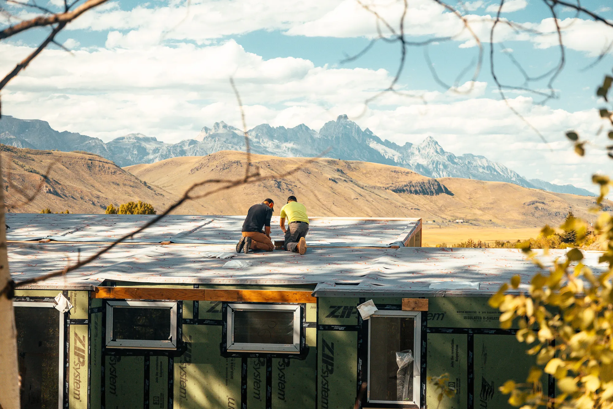 Two construction workers kneeling on a roof of a building under construction with mountains in the background.