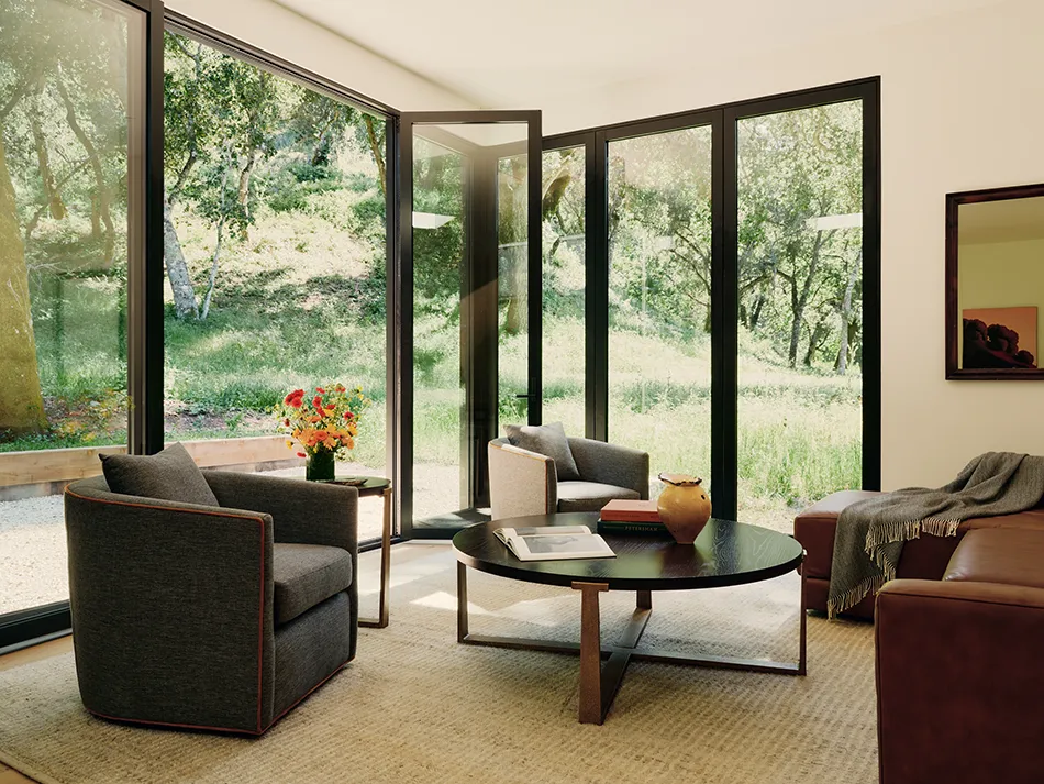Cozy living room with large glass doors opening to a green outdoor area, featuring armchairs and a coffee table.