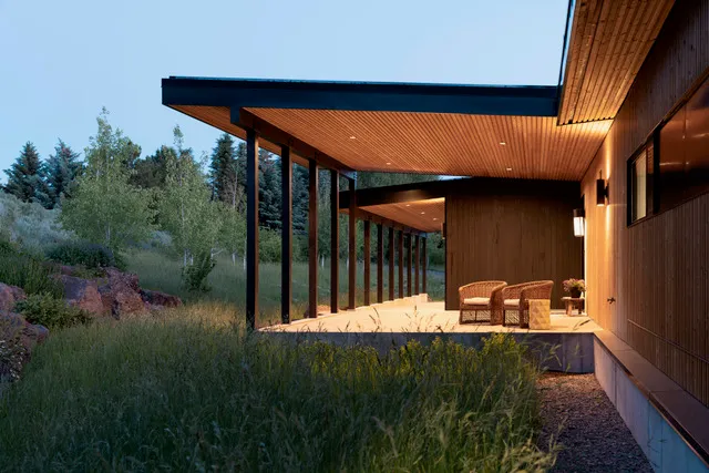 Modern wooden house porch with seating overlooking a grassy, wooded landscape at dusk.